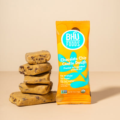 An individually wrapped Chocolate Chip Cookie Dough Premier Refrigerated Protein Bar with unwrapped cookie dough bars stacked beside it.