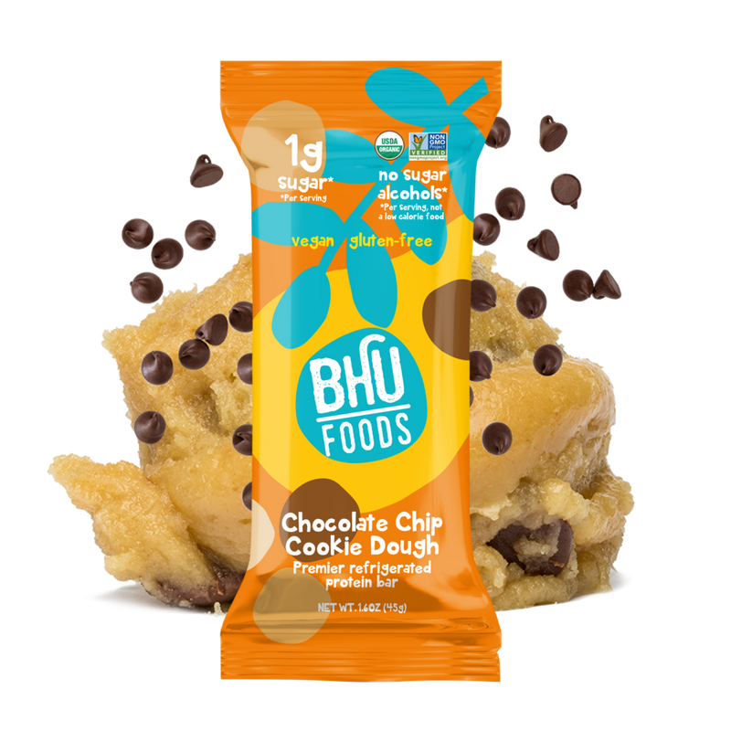 An individually wrapped Chocolate Chip Cookie Dough Premier Refrigerated Protein Bar with some chocolate chips and cookie dough behind it.	