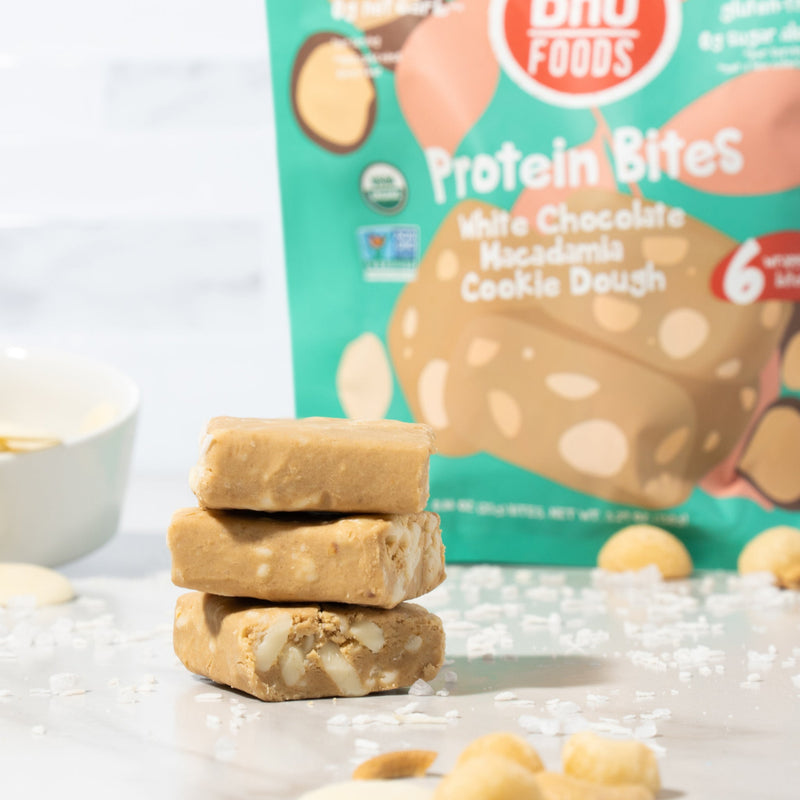 A close-up image of three unwrapped White Chocolate Macadamia Cookie Dough bites with some macadamia nuts and sea salt around and a bag of protein bites behind.