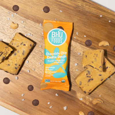 An individually wrapped Chocolate Chip Cookie Dough Premier Refrigerated Protein Bar with some chocolate chips, cashews, sea salt and unwrapped chocolate chip cookie dough around it.