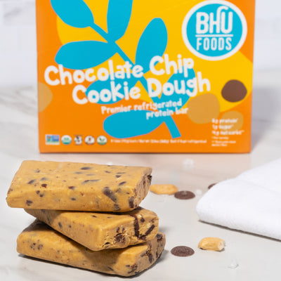 A closed box of Chocolate Chip Cookie Dough Premier Refrigerated Protein Bar with some chocolate chips, cashews, sea salt and three unwrapped chocolate chip cookie dough bars in front of the box.