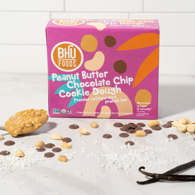 A closed box of Peanut Butter Chocolate Chip Cookie Dough Premier Refrigerated Protein Bar with some chocolate chips, peanuts, sea salt and a spoonful of peanut butter in front of the box.