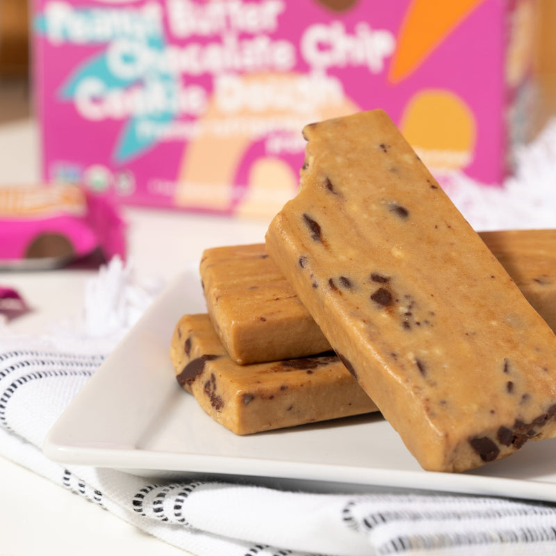 A close-up image of three unwrapped one partially bitten Peanut Butter Chocolate Chip Cookie Dough bars with a closed box of cookie dough bars behind.