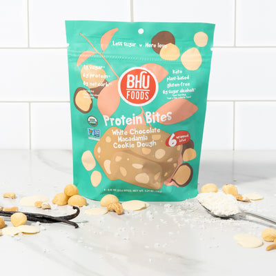 A bag of White Chocolate Macadamia Cookie Dough Protein Bites with some white chocolates, macadamia nuts and sea salt in front of it.
