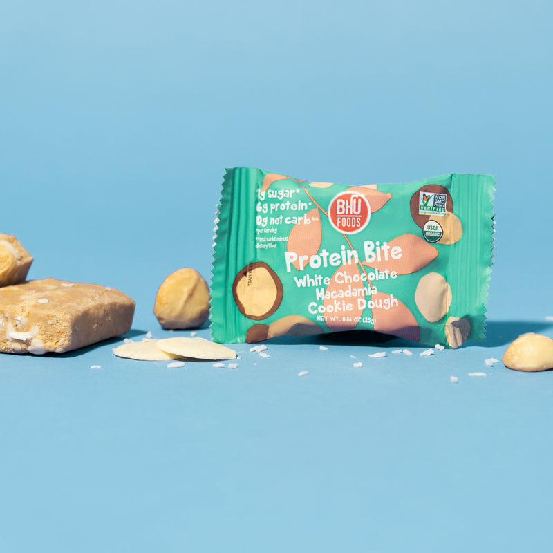 An individually wrapped White Chocolate Macadamia Cookie Dough Protein Bite with an unwrapped bite and some white chocolates, macadamia nuts and sea salt around it.