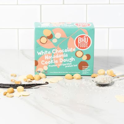 A closed box of White Chocolate Macadamia Cookie Dough Premier Refrigerated Protein Bar with some white chocolates, macadamia nuts and sea salt in front of the box.