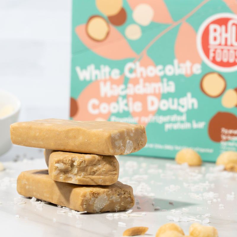 A close-up image of three unwrapped White Chocolate Macadamia Cookie Dough bars with some macadamia nuts and sea salt around and a closed box of cookie dough bar behind.
