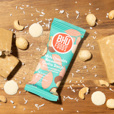 An individually wrapped White Chocolate Macadamia Cookie Dough Premier Refrigerated Protein Bar with some white chocolates, cashews, macadamia nuts, sea salt and unwrapped bars around it.