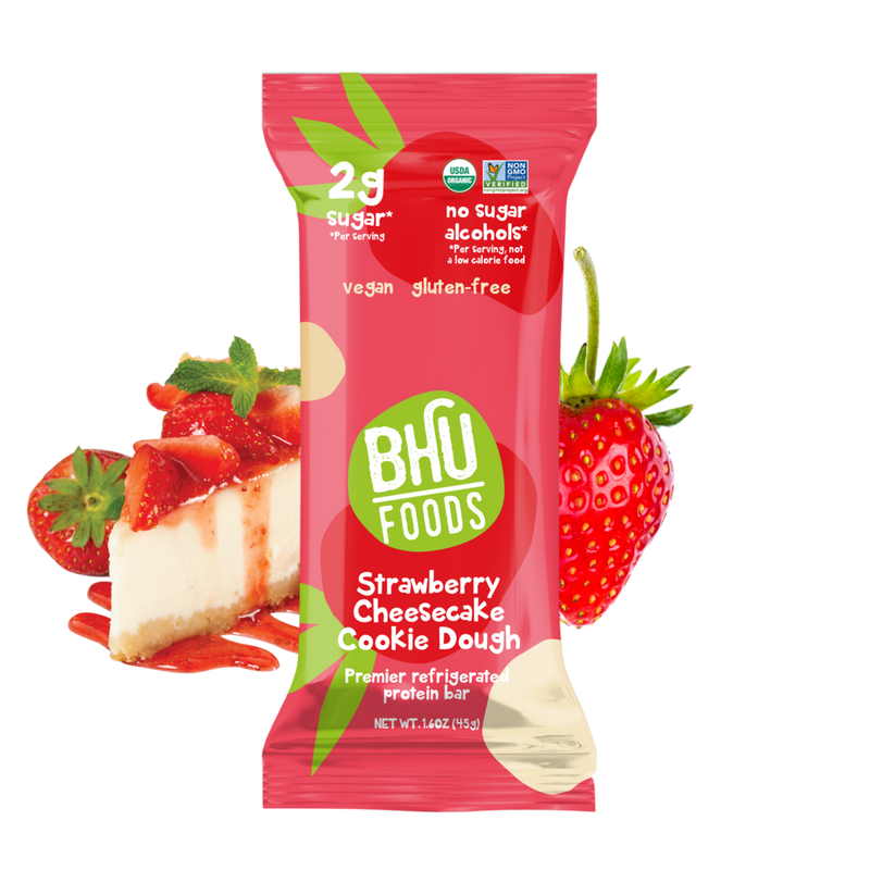 An individually wrapped Strawberry Cheesecake Cookie Dough Premier Refrigerated Protein Bar with a cheesecake and some strawberries behind it.	